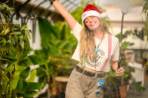 Lauren Davis of Fiddy Shades of Green stands in the greenhouse with one arm raised while wearing a Santa hat