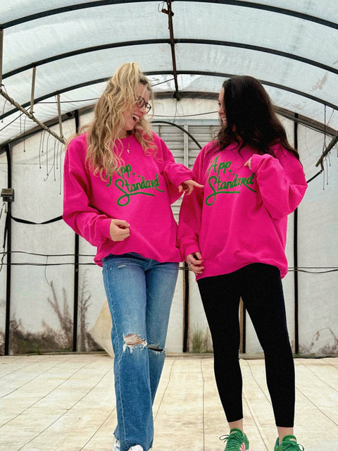 Lauren and Gracie standing wearing Appalachian Standard's pink and green sweatshirts in a white greenhouse.