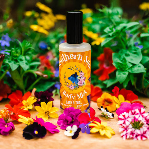 A bottle of Appalachian Standard's Southern Sun Hydrating Body Mist with Aloe on a piece of wood surrounded by flowers