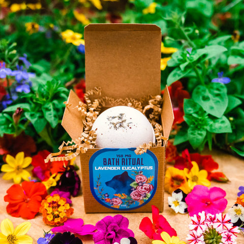 Lavender Eucalyptus Bath Bomb inside packaging on a table infront of plants surrounded by pink and red flowers by Appalachian Standard.