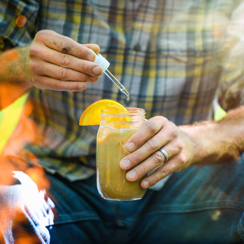 A man in a plaid tshirt and jeans dropping hemp tincture into a mason jar of orange chai.