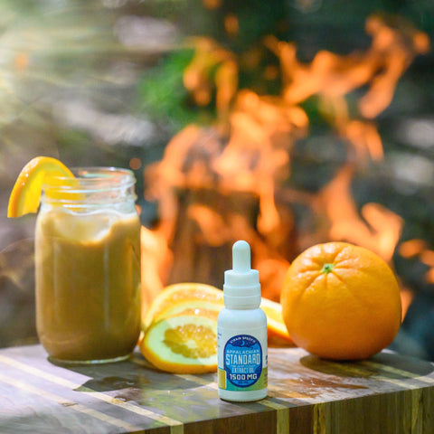 A bottle of Appalachian Standard's Orange Chai Hemp Oil Tincture on a wooden table with a mason jar of Orange Chai Latte and orange slices behind it.