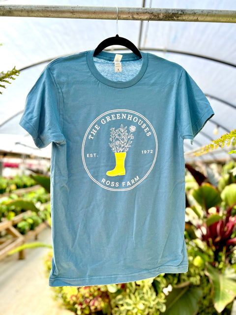 A close-up view of the same light blue Ross Farm Greenhouses t-shirt hanging from a metal pipe in a greenhouse structure. The shirt displays the circular logo with the yellow boot filled with illustrated flowers and plants.