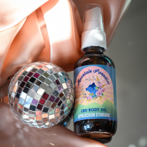 A small disco ball next to a amber glass bottle of Mountain Paradise CBD Body Oil by Appalachian Standard. The bottle features a colorful sunset label with mountain silhouettes, floral designs, and a the bath rituals product line bear logo.