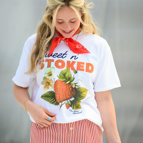 A person with long blonde hair looking down with a smile, wearing a white "Sweet n' Stoked" t-shirt featuring a detailed botanical illustration of a strawberry with leaves and flowers. The person has a red bandana tied around their neck, turquoise rings, and is wearing red and white striped shorts. The brand name "Appalachian Standard" appears at the bottom of the design.
