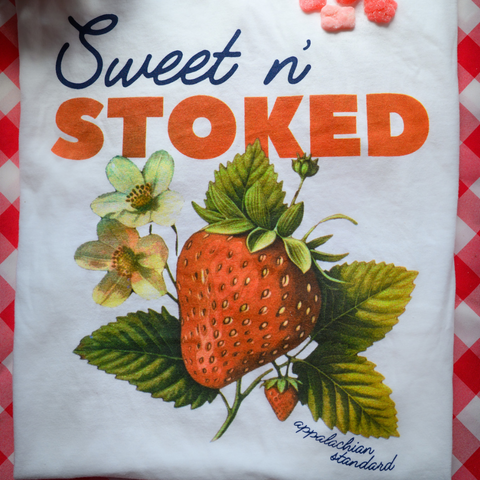A close-up detail shot of the "Sweet n' Stoked" t-shirt graphic showing the botanical strawberry illustration against a white background with a red checkered border. The design features detailed strawberry fruit with green leaves and delicate strawberry blossoms. Some pink strawberry-shaped gummies are visible at the top of the image. The "Appalachian Standard" logo appears at the bottom of the design.