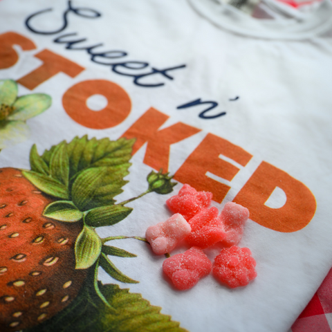 An extreme close-up of the "Sweet n' Stoked" t-shirt design focusing on the brand name in orange letters and part of the strawberry illustration. Several pink strawberry-shaped gummies are placed on the shirt, showing both the product and the matching graphic design theme.