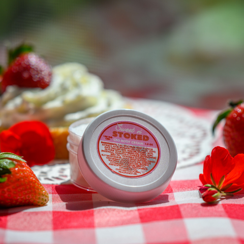 A small white container of Sweet n' Stoked CBD body cream (1.5 oz) on a red and white checkered tablecloth, surrounded by strawberries and red flowers. The product label indicates it contains 188 mg of CBD and lists various natural ingredients.