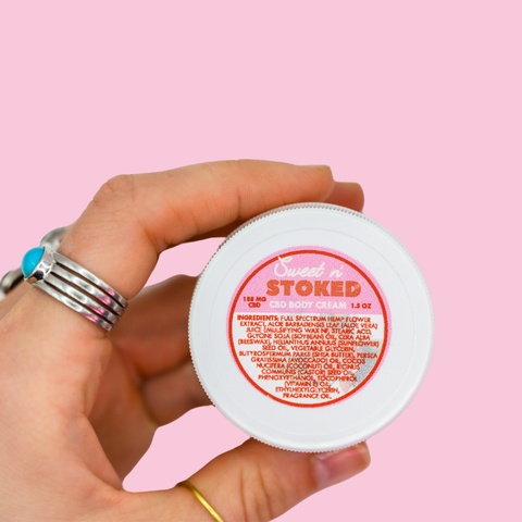 A hand holding the Sweet n' Stoked CBD body cream against a pink background, providing scale and showing the same silver ring with turquoise stone. The product's ingredient list is visible on the label.