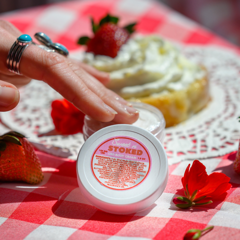 A close-up view of someone's hand with turquoise jewelry holding or pointing to the same Sweet n' Stoked CBD body cream. The product is placed on what appears to be a decorative plate with a dessert topped with whipped cream and a strawberry in the background, still on the red and white checkered tablecloth.