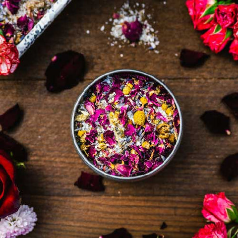 An 8 oz tin filled with bath salts, rose petals, chamomile flowers, and lavender from Appalachian Standard's CBD Botanical Dream Bath Soak on a piece of wood surrounded by flowers