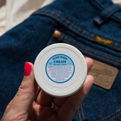 Appalachian Standard Dad Bod Cream, 1.5 oz jar, with 188mg CBD, being held by a hand over a pair of Wrangler jeans.