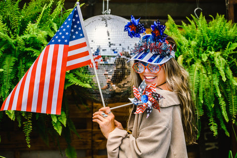 Person holding an American flag and festive hat with a disco ball and ferns in the background