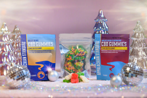 A photo of Appalachian Standards Yuletide Yummy Gummies featuring Limited Edition Christmas Boujee Bears and Boujee Bites, paired with their best-selling Classic Boujee Bears and Elderberry CBD Gummies. The Gummies are surrounded by shiny glass Christmas trees, disco balls, and ornaments.