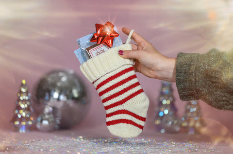 A photo of Appalachian Standards Kris Kringles' CBD Chocolate Stocking. This bundle includes their Limited Edition Teeny Weeny Marshmallow CBD Chocolate, Raspberry Almond, Salted Croissant, and Caramel Macchiato. The Chocolate bars are placed in a white and red striped stocking with shiny glass christmas tree decorations, disco balls, and ornaments in the background.