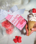 Hand holding a ice cream cone with raspberry toppings next to a bag of red candies and a pink label.