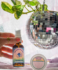 Appalachian Standard's Mountain Paradise CBD Body Oil being held by a hand with a gold ring, and CBD Body Cream on a white table with a purple drape and hanging disco ball planter with a philodendron inside.