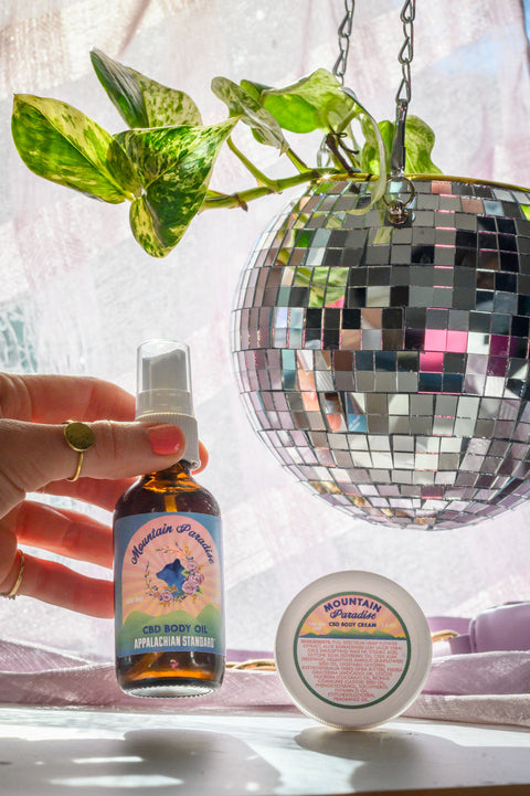 Appalachian Standard's Mountain Paradise CBD Body Oil being held by a hand with a gold ring, and CBD Body Cream on a white table with a purple drape and hanging disco ball planter with a philodendron inside.