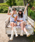 Two young women sitting on the tailgate of a white pickup truck, wearing matching white t-shirts and baseball caps. They're holding up small packages while smiling at the camera. The scene is set near a pond with lush green vegetation in the background.
