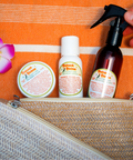 Appalachian Standard Coconut Lime Redneck Riviera CBD skincare set — body cream, body lotion, and after-sun mist — displayed in the open dark woven straw travel bag on an orange beach towel with a tropical flower