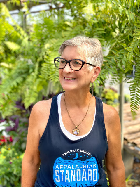 Woman wearing a blue tank top with 'Appalachian Standard' text, standing outdoors with greenery in the background.