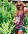 Woman standing among tall green plants wearing a purple tank top with visible branding.