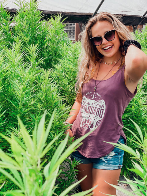 Woman standing among tall green plants wearing a purple tank top with visible branding.
