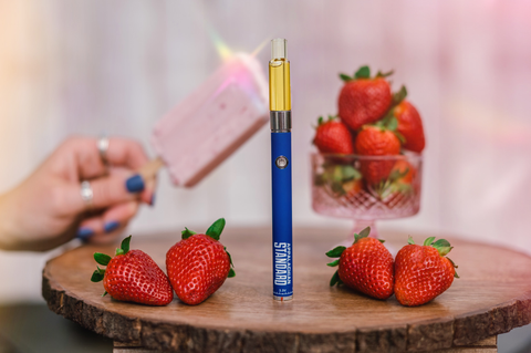 A styled product photo of a blue Appalachian Standard vape pen from Asheville, NC, labeled with 'APPALACHIAN STANDARD' and '3.3V' standing upright on a round wooden slice. The device offers calming plant goodness. It's surrounded by fresh red strawberries. In the blurred background, there's a crystal cup filled with more strawberries and a hand holding what appears to be a pink popsicle or ice cream bar. The hand has blue nail polish and is wearing rings. The background has a soft pink tone.