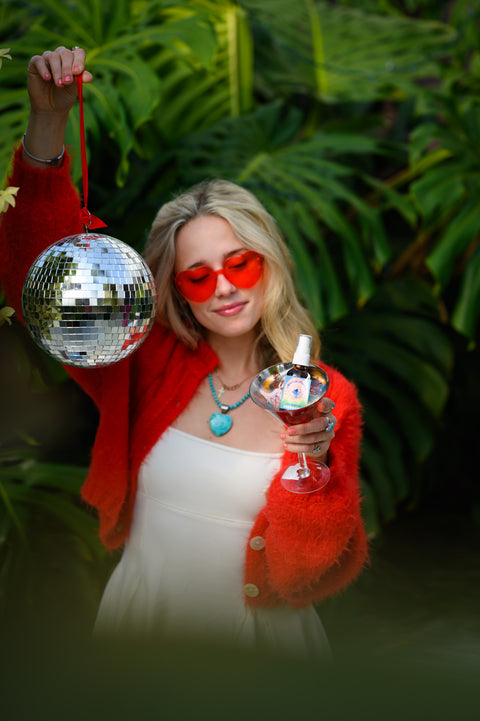 Appalachian Standard's Mountain Paradise CBD Body Oil in a martini glass being held by a blonde girl in a red sweater, holding a disco ball in front of a big green monstera plant.