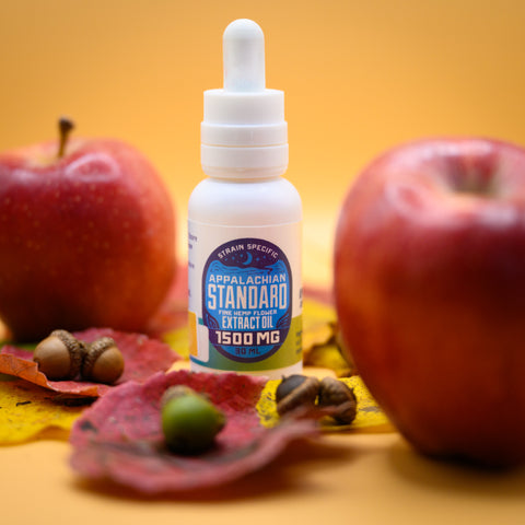 Appalachian Standard's CBD Apple-achian Tincture, in from of an orange background, surrounded by res apples, fall leaves, and acorns.