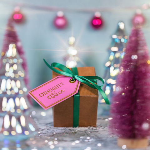 Gift box with a pink tag labeled 'Naughty or Nice' against a festive background with Christmas trees and lights.