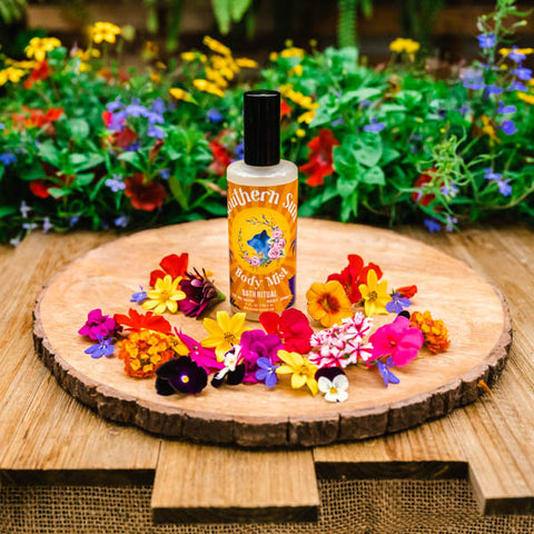 A bottle of Appalachian Standard's Southern Sun Hydrating Body Mist with Aloe on a round wood platter surrounded by flowers