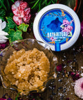 An 8 oz tin of Appalachian Standard's CBD Body Polish and brown sugar in a glass jar on a piece of wood surrounded by flowers and bath salts