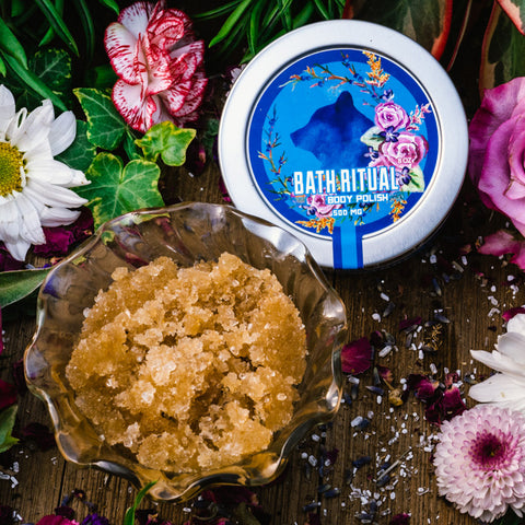 An 8 oz tin of Appalachian Standard's CBD Body Polish and brown sugar in a glass jar on a piece of wood surrounded by flowers and bath salts