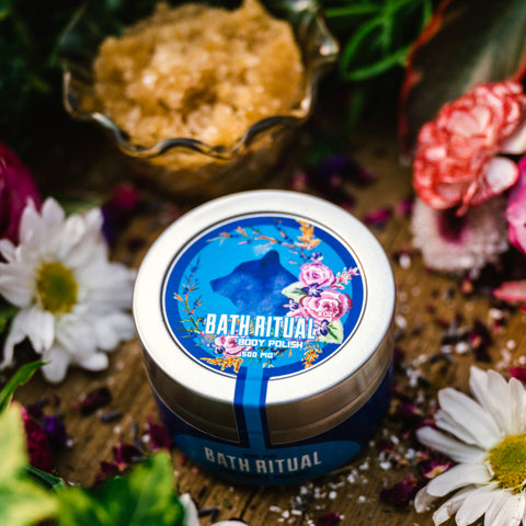 An 8 oz tin of Appalachian Standard's CBD Body Polish on a piece of wood surrounded by flowers, bath salts, and a glass jar of brown sugar