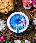 An 8 oz tin of Appalachian Standard's CBD Body Polish on a piece of wood surrounded by flowers, bath salts, and brown sugar