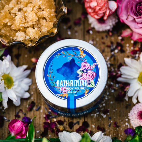 An 8 oz tin of Appalachian Standard's CBD Body Polish on a piece of wood surrounded by flowers, bath salts, and brown sugar