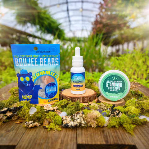 The three products from Appalachian Standard's CBD Starter Kit Bundle: a 1 oz bottle of the Balance Tincture, a bag of Boujee Bears, and a tin of CBD Salve on wood slices surrounded by moss with plants in the background