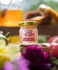 Jar of Appalachian Standard High Country Hunny 15mg CBD-infused honey, 2oz, collaboration with HoneyBeeBliss.com, styled with apples, a pear, and purple and yellow flowers, with a honey dispenser and pastries blurred in the background.