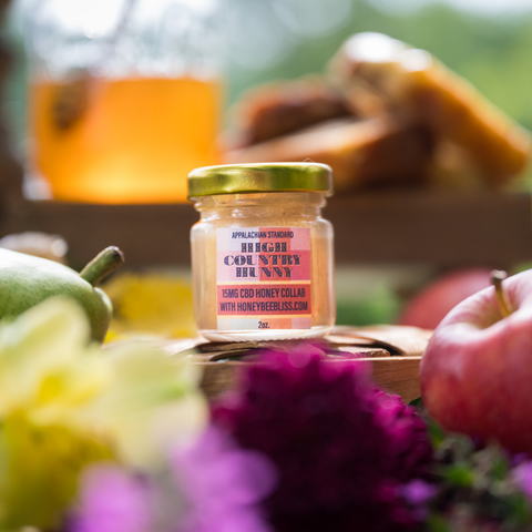Jar of Appalachian Standard High Country Hunny 15mg CBD-infused honey, 2oz, collaboration with HoneyBeeBliss.com, styled with apples, a pear, and purple and yellow flowers, with a honey dispenser and pastries blurred in the background.