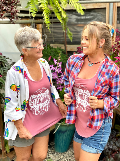 Two women standing in the greenhouse in dusty rose Appalachian Standard tank tops looking at each other