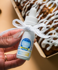 Hand holding a small white bottle with a blue label in front of a cinnamon roll.