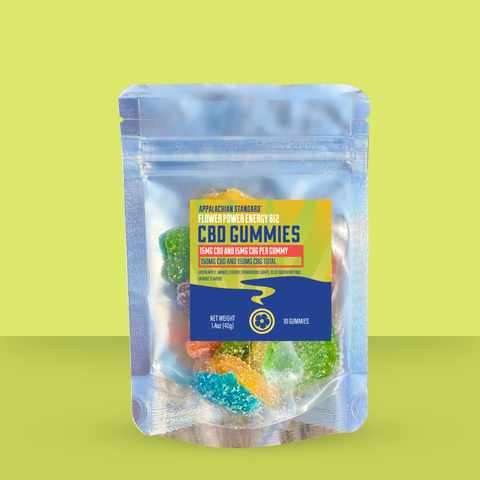 Appalachian Standard Flower Power Energy B12 CBD Gummies in a clear resealable mylar pouch against a lime green background. The label displays 15mg CBD and 15mg CBG per gummy, 150mg total, in green apple, mango, cherry, strawberry, grape, blue raspberry, and orange flavors. Net weight 1.4oz (40g), 10 gummies. Colorful sour gummy candies are visible through the packaging.