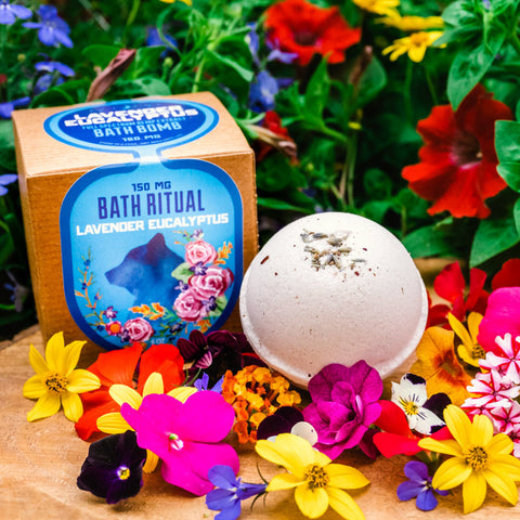 Appalachian Standard's Lavender Eucalyptus CBD Bath Bomb next to its box on a piece of wood surrounded by flowers