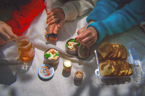 A cozy overhead scene of hands sharing small jars of spreads and snacks with sliced bread on a blanket. Visible colors include a bright blue sleeve, a cream knit sleeve, a red sleeve, and the white blanket underneath.