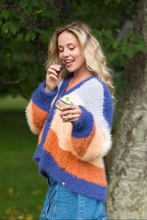 A woman stands outdoors in a park-like setting with trees and fallen leaves, wearing a chunky knit cardigan with blue, orange, and cream stripes, a denim skirt, and brown boots.