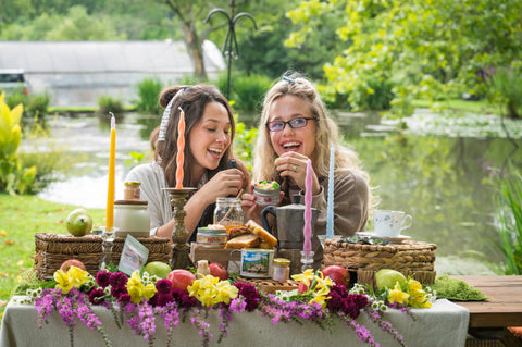 Lauren and Gracie laughing together at an outdoor table styled with High Country Hunny products, honey jars, flowers, fruit, and a picnic spread by a pond