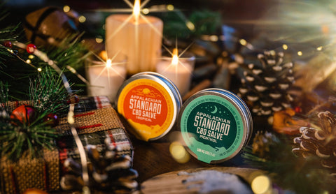 Appalachian Standard's CBD Salve in front of a Christmas setting of pinecones, presents, candles and bells.