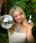 Girl in white dress holding disco ball and amber bottle of Mountain Paradise Body Oil up with plants blurred in background