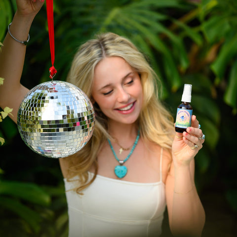 Girl in white dress holding disco ball and amber bottle of Mountain Paradise Body Oil up with plants blurred in background